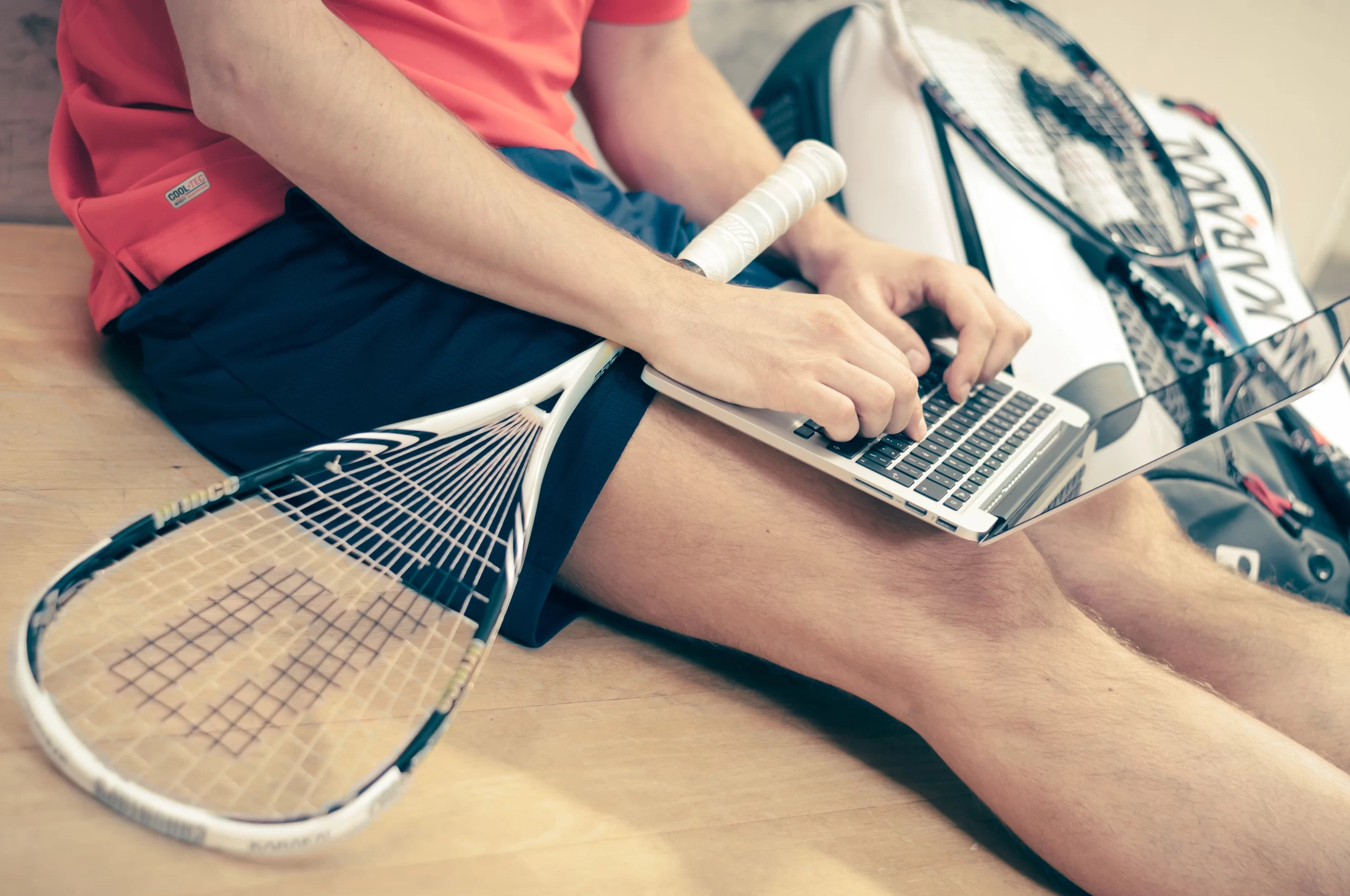 A young tennis athlete sitting ,holds a tennis racket and uses his laptop on his lap.