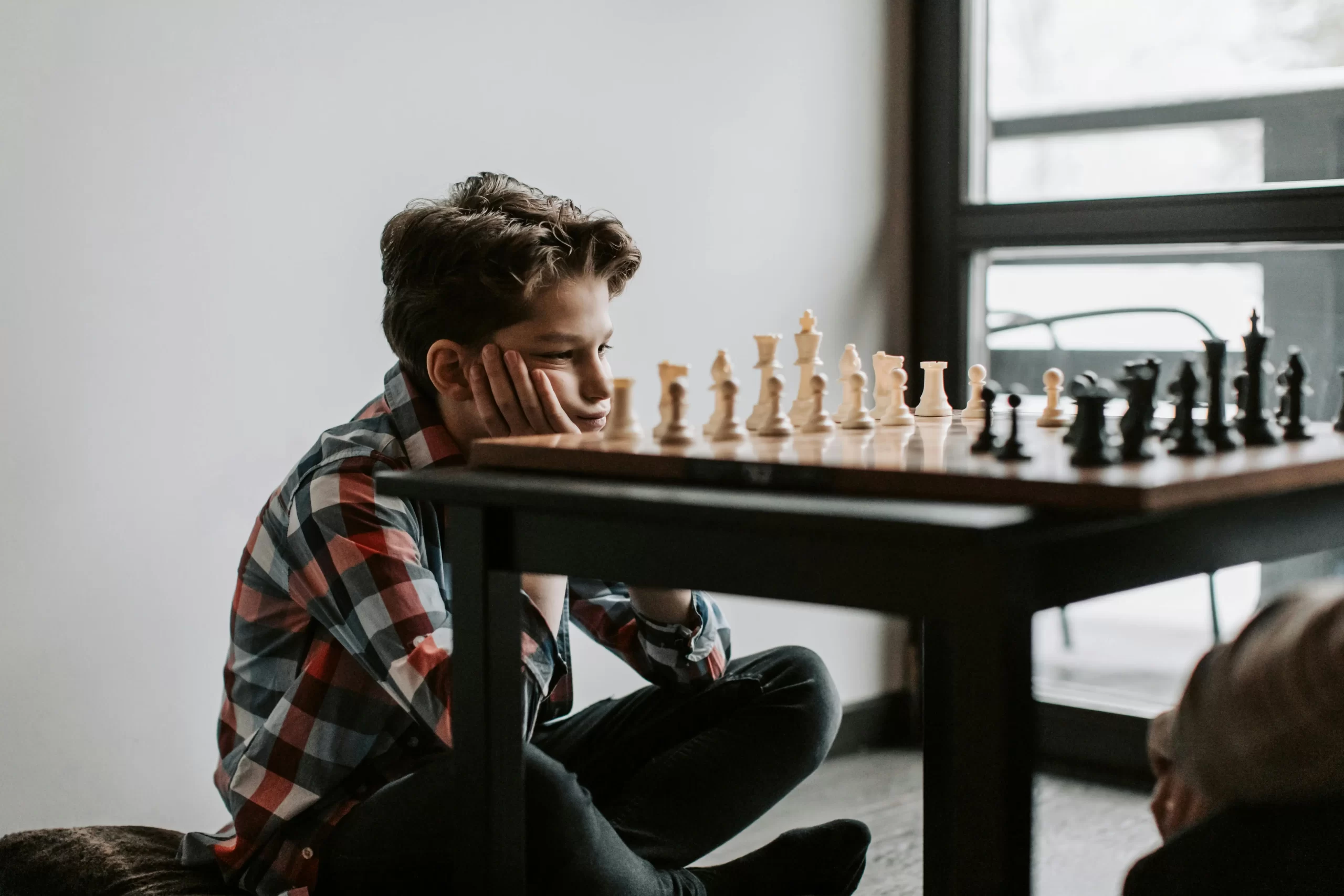 A young boy in a plaid shirt sits cross-legged on the floor, intently contemplating his next move during a chess game.